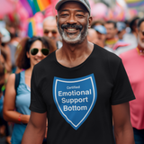 A smiling man wearing a black t-shirt with a blue shield graphic that reads 'Certified Emotional Support Bottom' stands in a crowded outdoor street scene with rainbow flags in the background.