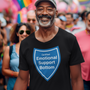 A smiling man wearing a black t-shirt with a blue shield graphic that reads 'Certified Emotional Support Bottom' stands in a crowded outdoor street scene with rainbow flags in the background.