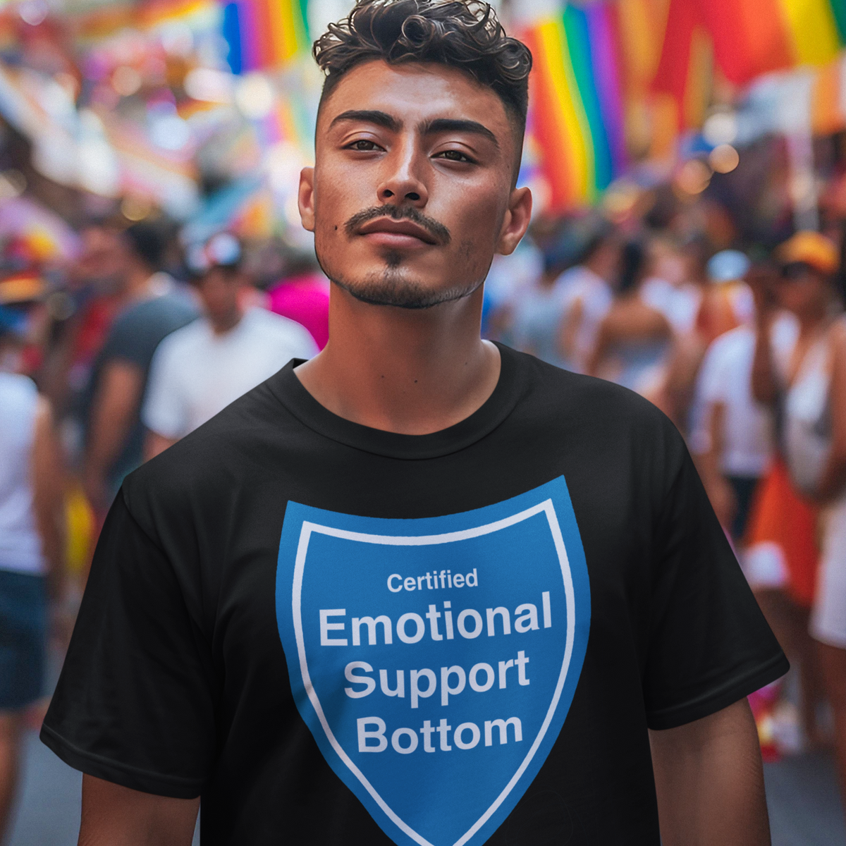 A man wearing a black t-shirt with a blue shield graphic that reads 'Certified Emotional Support Bottom' stands in a crowded street during a pride parade with rainbow flags in the background.
