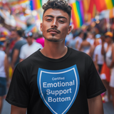 A man wearing a black t-shirt with a blue shield graphic that reads 'Certified Emotional Support Bottom' stands in a crowded street during a pride parade with rainbow flags in the background.