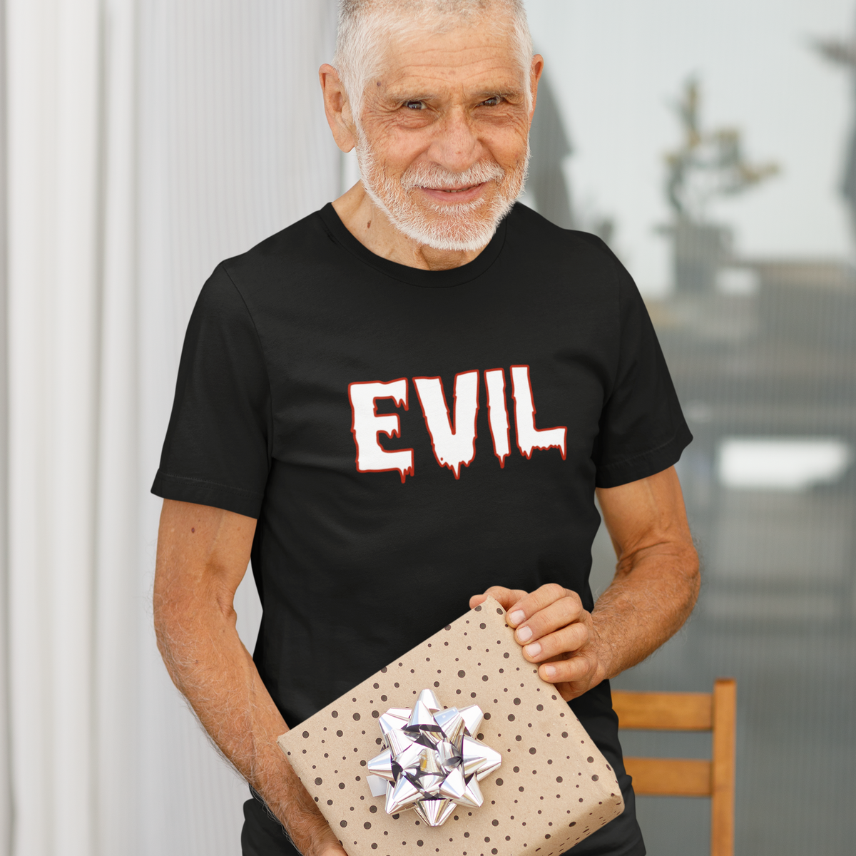 An older man with a white beard stands behind a dining table holding a wrapped gift box. He is wearing a black t-shirt with the word 'EVIL' printed in a dripping red-and-white font.