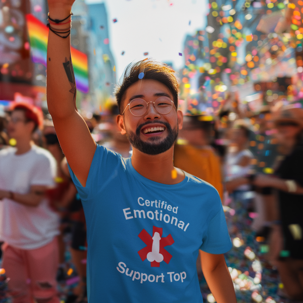 A smiling man wearing glasses and a blue graphic T-shirt waves his hand in a crowded outdoor street scene filled with confetti and rainbow flags. The shirt reads 'Certified Emotional Support Top' with a red heart and white ribbon graphic.