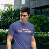 A man wearing a slate blue crewneck t-shirt with the text 'THANKS FOR LEAVING!' printed in orange and white on the chest, standing on an outdoor lawn with green plants and a modern building in the background.